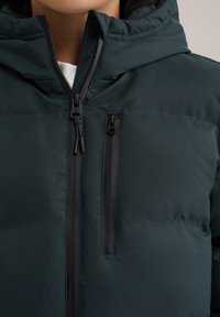 Temnozelena puhovka z zadrgo na sprednji strani, opremljena z črnim žepom na prsih z zadrgo, gladko teksturo in nastavljivo kapuco.