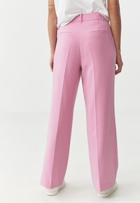 Roze wijde broek met een zachte textuur, twee achterzakken en gestikte details, gecombineerd met witte sneakers op een neutrale achtergrond.