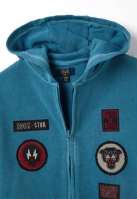 Sudadera con capucha azul que presenta un cierre de cremallera, tela de algodón texturizada y múltiples parches bordados con diseños y colores variados.