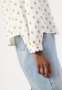 Vit blus med svart geometriskt mönster, puffärmar, elastiska manchetter och scallop-kant, i kombination med ljusblå denimjeans.