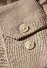 Beige texturerat tyg med två ljusa knappar på en krage, som visar ett subtilt vävt mönster och en ren, minimalistisk design.