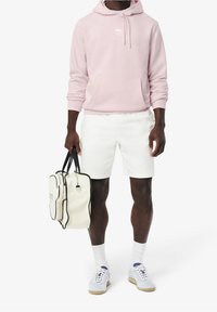 Světle růžová mikina s kapucí a přední kapsou, bílé šortky a bílá taška s černými doplňky. Bílé tenisky s modrými detaily podtrhují neformální vzhled.