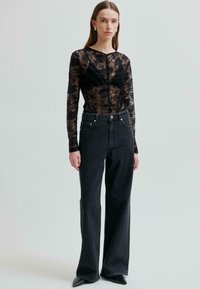 Młoda kobieta w prześwitującej czarnej koronkowej bluzce z długim rękawem, czarnym biustonoszu, czarnych jeansach z wysokim stanem i czarnych czółenkach, stojąca na tle bez wzorów.