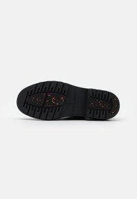 Semelle de chaussure en caoutchouc noir avec un motif de grip texturé, présentant des éclaboussures rouges au centre et un logo en relief dans la zone du talon.