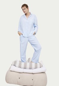 Lichtblauw en wit gestreept pyjama-set met een knoopsluiting shirt en bijpassende broek, tentoongesteld op een gelaagde, gestoffeerde stoffen platform.