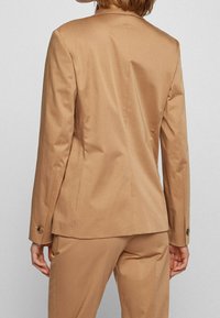 Widok z tyłu osoby w dopasowanej czarnej marynarce w kolorze wielbłądzim i pasujących spodniach na tle jednolitego jasnego tła.