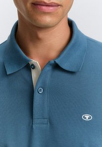 Camisa polo azul con tejido texturizado; presenta un tapeta blanca y dos botones, acentuada por un pequeño logo en el pecho.