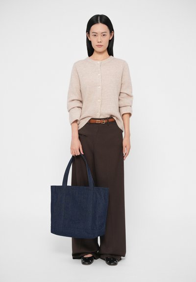 Τσάντα tote από ναυτικό τζιν με δύο λαβές, σταθερή βάση και διακριτική λεπτομέρεια κειμένου. Συνδυασμένη με ανοιχτό μπεζ ζακέτα και καφέ παντελόνια.