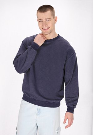 Junger Mann mit kurzen Haaren, der lächelt, ein navy-blaues Sweatshirt und eine hellblaue Cargo-Hose trägt und mit einer Hand nahe seinem Hals posiert.