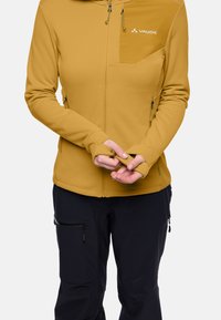 Giacca gialla senape con zip e fori per il pollice, tasca sul petto e cerniere. Abbinata a pantaloni scuri con tasche e cerniere.