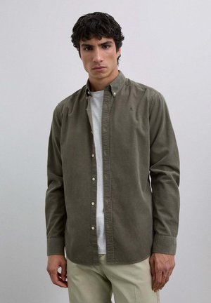 Camisa de botón color caqui en suave tejido de algodón, con collar clásico y corte ajustado. Usada sobre una camiseta blanca y combinada con pantalones claros.