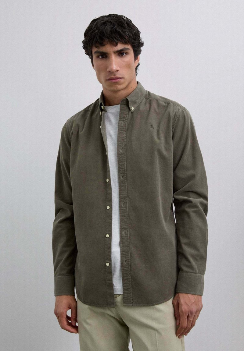 Khaki button-up shirt in soft cotton fabric, featuring a classic collar and a slim fit. Worn over a white t-shirt and paired with light pants.