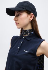 Νεαρή γυναίκα που φοράει μπλε καπέλο και αμάνικο πόλο με το λογότυπο Barbour, φλοράλ μαντήλι στο λαιμό και μικρά σκουλαρίκια κρίκους, κοιτάζοντας πλάγια.