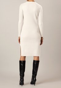 Robe tricotée blanche longue, à côtes, avec des manches longues et des fentes sur les côtés, associée à des bottes noires à talons hauts jusqu'aux genoux.