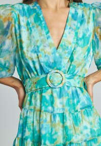 Un vestito turchese con scollatura a V, maniche a sbuffo e design a strati. Presenta una cintura larga con una fibbia circolare e dettagli in oro.