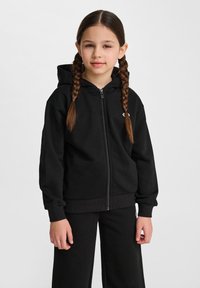 Czarna bluza na zamek, wykonana z miękkiego materiału, z kapturem, ściągaczami na mankietach oraz małym logo na piersi. Noszona z pasującymi spodniami.