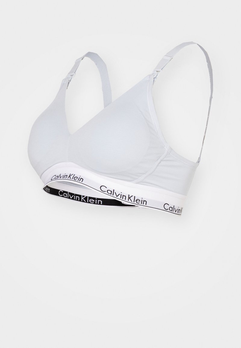 Gaišpelēks mīksts krūšturis ar regulējamām siksnām un plašu elastīgu lentu, kurai ir kontrastējošs melnbalts Calvin Klein logotips.