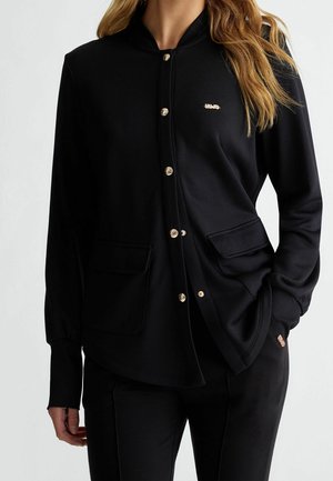 Femme portant une veste noire boutonnée avec deux poches avant et un petit logo doré en haut à gauche.