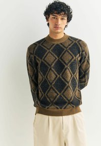 Maglione lavorato a maglia con un motivo a diamante testurizzato in marrone e blu navy, con un colletto a girocollo a coste e orlo. Indossato sopra pantaloni chiari.