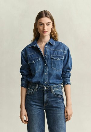 Vrouw met lang, golvend haar, gekleed in een blauw denim overhemd met knopen in een blauwe spijkerbroek tegen een effen lichte achtergrond.