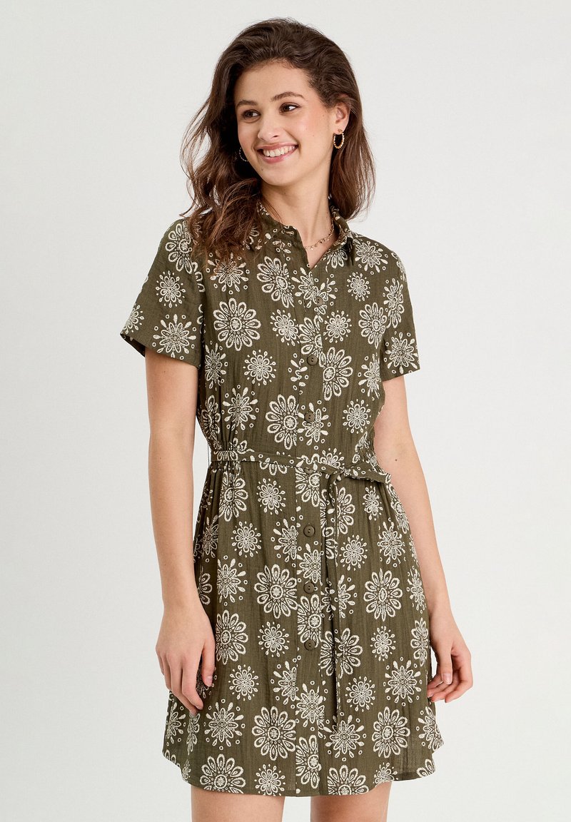 Robe verte à manches courtes avec un devant à boutons, motif floral blanc sur l'ensemble, ceinture à nouer à la taille et tissu texturé.