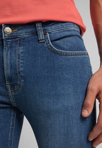 Pantalones de mezclilla de color azul oscuro, corte recto, con una textura sutil y costuras amarillas. Presentan un botón de la marca Lee y un bolsillo delantero.
