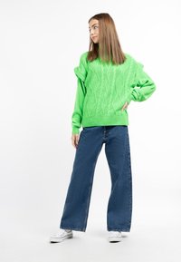 Pulover verde tricotat cu un design texturat de cabluri, asortat cu blugi evazați albaștri și adidași albi.