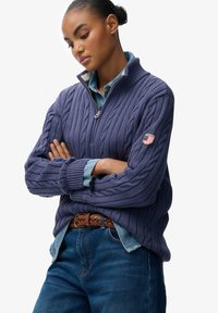 Marineblauer Kabelstrickpullover mit Reißverschlusskragen, kombiniert mit einem Denimhemd. Verfügt über ein kleines gesticktes Logo am Ärmel und ein strukturiertes Design.