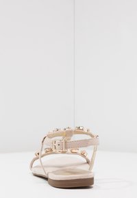 Beige litte sandaler har et smalt bånddesign med en spænde-closure, prydet med juvel-accenter langs stropperne og en tekstureret fodseng.