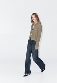 Khaki sweterkowy kardigan z prążkowanym splotem i kapturem, czarne guziki, zestawiony z ciemnoniebieskimi szerokimi dżinsami i czarnymi szpilkami w szpic.