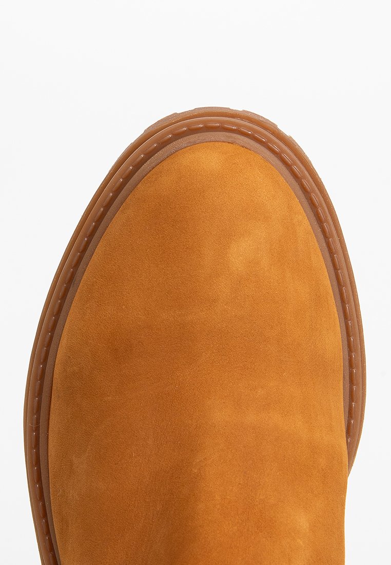 Botín de ante beige con punta redonda, suela de goma contrastante y costuras visibles a lo largo del borde. Textura suave y acabado liso.