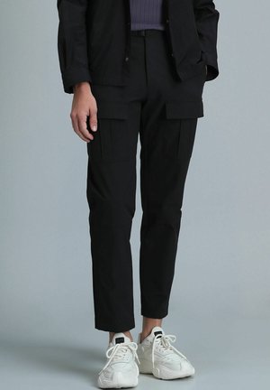 Pantalons cargo noirs avec une coupe fuselée, deux poches avant et une texture subtile. Associés à des baskets blanches, montrant des lacets et un design minimaliste.