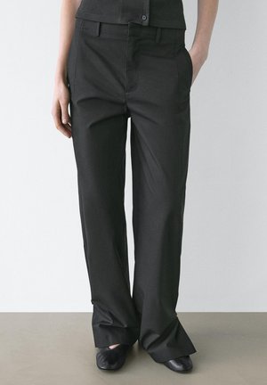 Person wearing loose black straight-leg pants with side pockets and black flat shoes, standing against a plain wall.