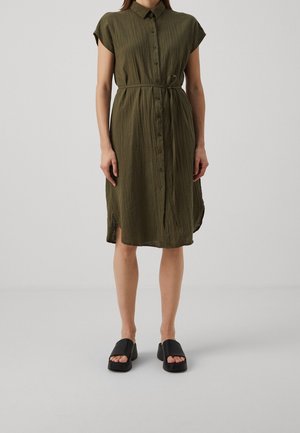 Femme portant une robe boutonnée mi-longue de couleur olive avec manches courtes, ceinture à la taille, et sandales à plateforme noires à bout ouvert, debout à l'intérieur.
