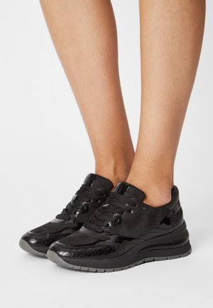 Jambes portant des baskets basses noires en cuir texturé avec des semelles épaisses grises, debout sur fond blanc.
