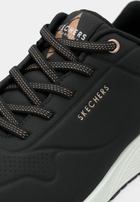 Μαύρο αθλητικό παπούτσι με υφή στην επάνω επιφάνεια, με καφέ κορδόνια ως διακοσμητικό στοιχείο, μεταλλικό λογότυπο και λευκή σόλα με ανάγλυφο πέλμα.