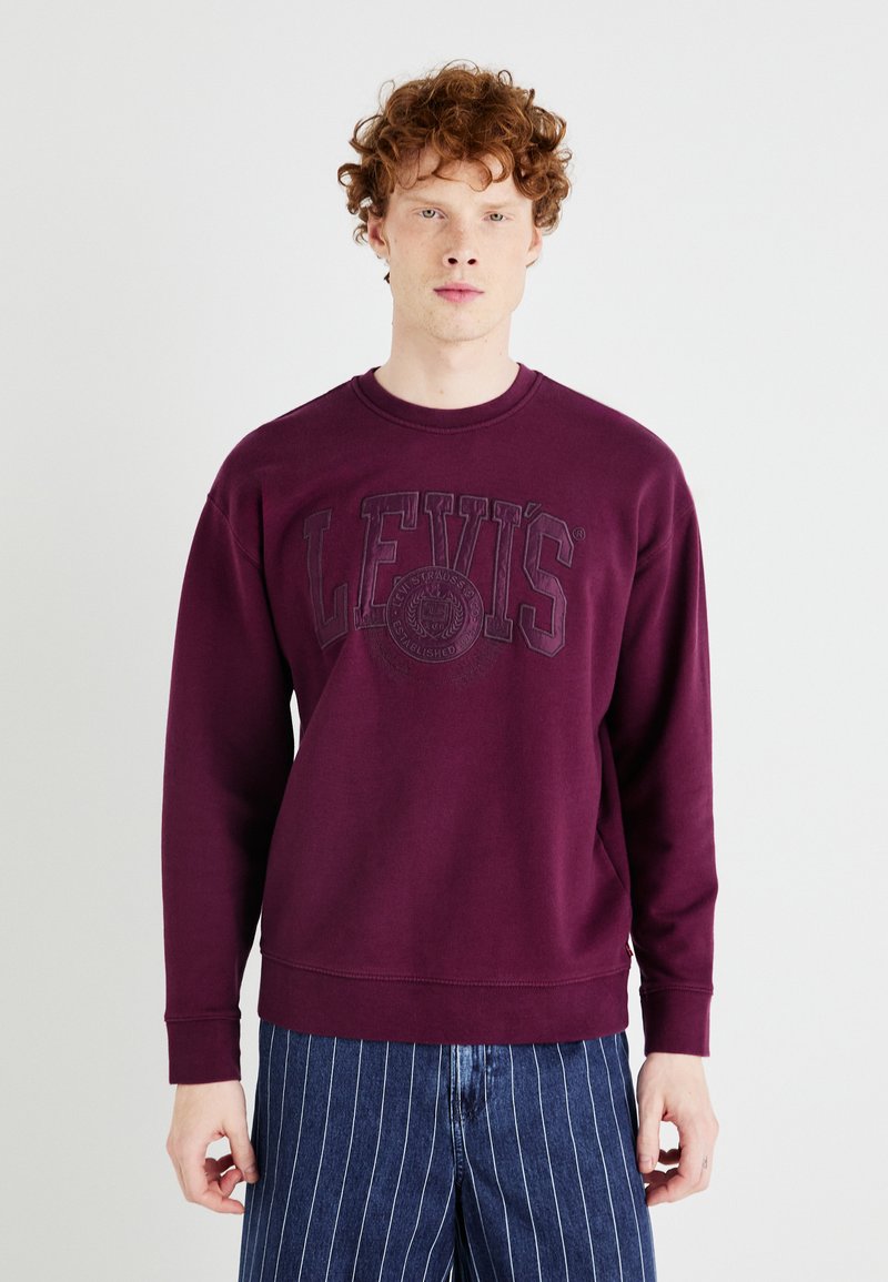 Felpa bordeaux con logo "LEVI'S" ricamato, colletto rotondo, maniche lunghe e polsini a coste; abbinata a pantaloni blu con righe sottili.