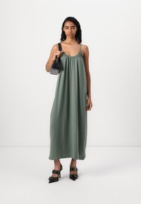 Green, sleeveless maxi dress made of lightweight fabric, featuring a gathered neckline. Complemented by black pointed shoes and a small handbag.
