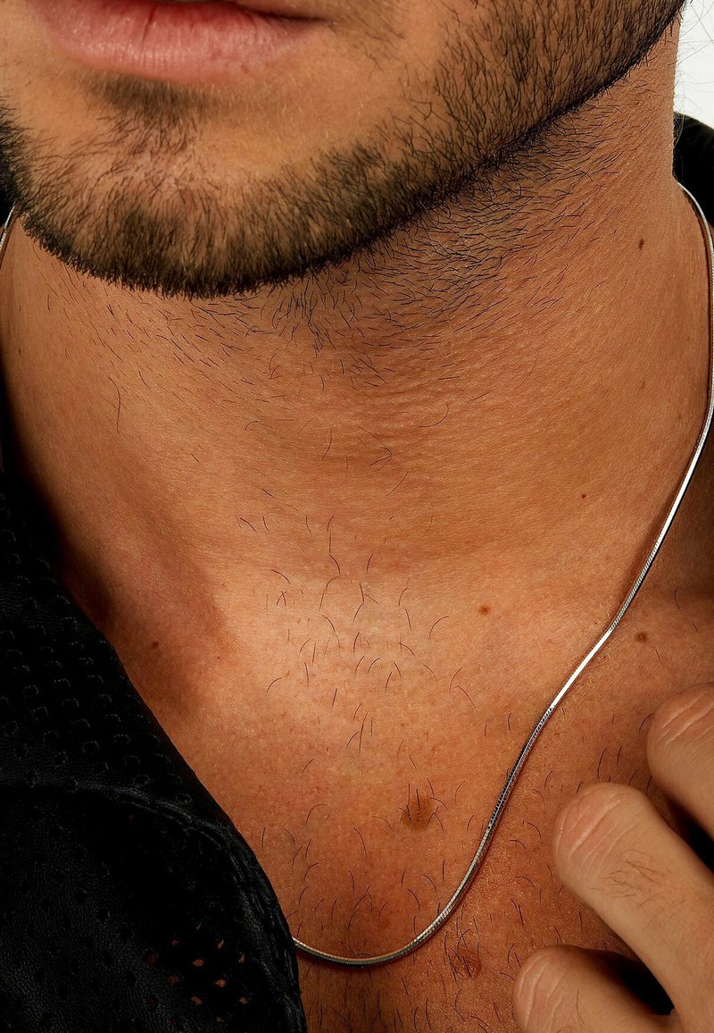 Silberne Halskette, die um den Hals gelegt ist, kontrastiert mit gebräunter Haut und feinem dunklen Haar. Schwarzer Shirtstoff sichtbar mit perforierter Textur.