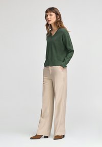 Pullover vert à col en V avec des manches longues, associé à un pantalon large beige et des bottines marron. Texture de tissu lisse tout au long.