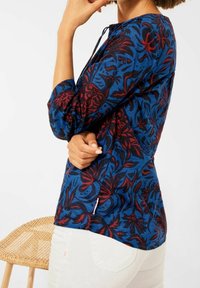 Blouse bleue à motif floral avec des accents rouges, dotée de manches trois-quarts, d'un col en trou de serrure et d'une légère fente latérale.