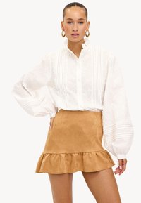 Witte blouse met lange mouwen, versierd met textuurpatronen en een hoge kraag, gecombineerd met een tan kleurige, suède mini rok met een gerimpelde zoom.