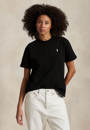 Femeie cu păr creț purtând o bluză neagră cu mânecă scurtă cu logo alb și pantaloni albi, stând în fața unui fundal neutru.