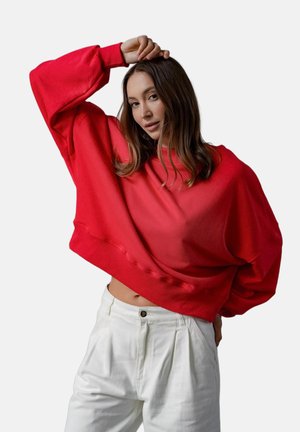 Kobieta o długich brązowych włosach, ubrane w luźny czerwony sweter i białe spodnie, pozująca z uniesionym jednym ramieniem, dłoń spoczywa na jej głowie.