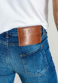 Los pantalones vaqueros de denim azul cuentan con una parche de cuero marrón con el texto en relieve "GIANNI KAVANAGH". La tela muestra un ligero desvanecimiento y detalles de costura.