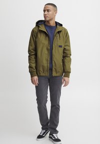 Olive green lightweight jacket with hood, elastic cuffs, and hem; paired with a navy sweater, gray jeans, and black-and-white sneakers.