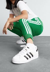 Vita Adidas Superstar-sneakers med svarta ränder, bärs med gröna träningsbyxor som har vita sidosträck och en kortärmad vit topp.
