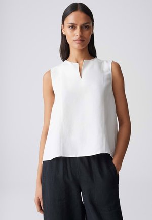 Femme aux longs cheveux foncés portant un haut blanc sans manches avec une fente à l'encolure devant et un pantalon noir, les mains dans les poches.