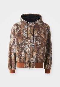 Camo zip-up hoodie in bruin en beige met bladerenprints, een trekkoordkap en contrasterende bruine manchetten. De stof lijkt textuur te hebben.