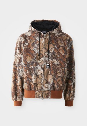 Camo zip-up hoodie in brown and beige with leaf prints, drawstring hood, and contrasting brown cuffs. Fabric appears textured.